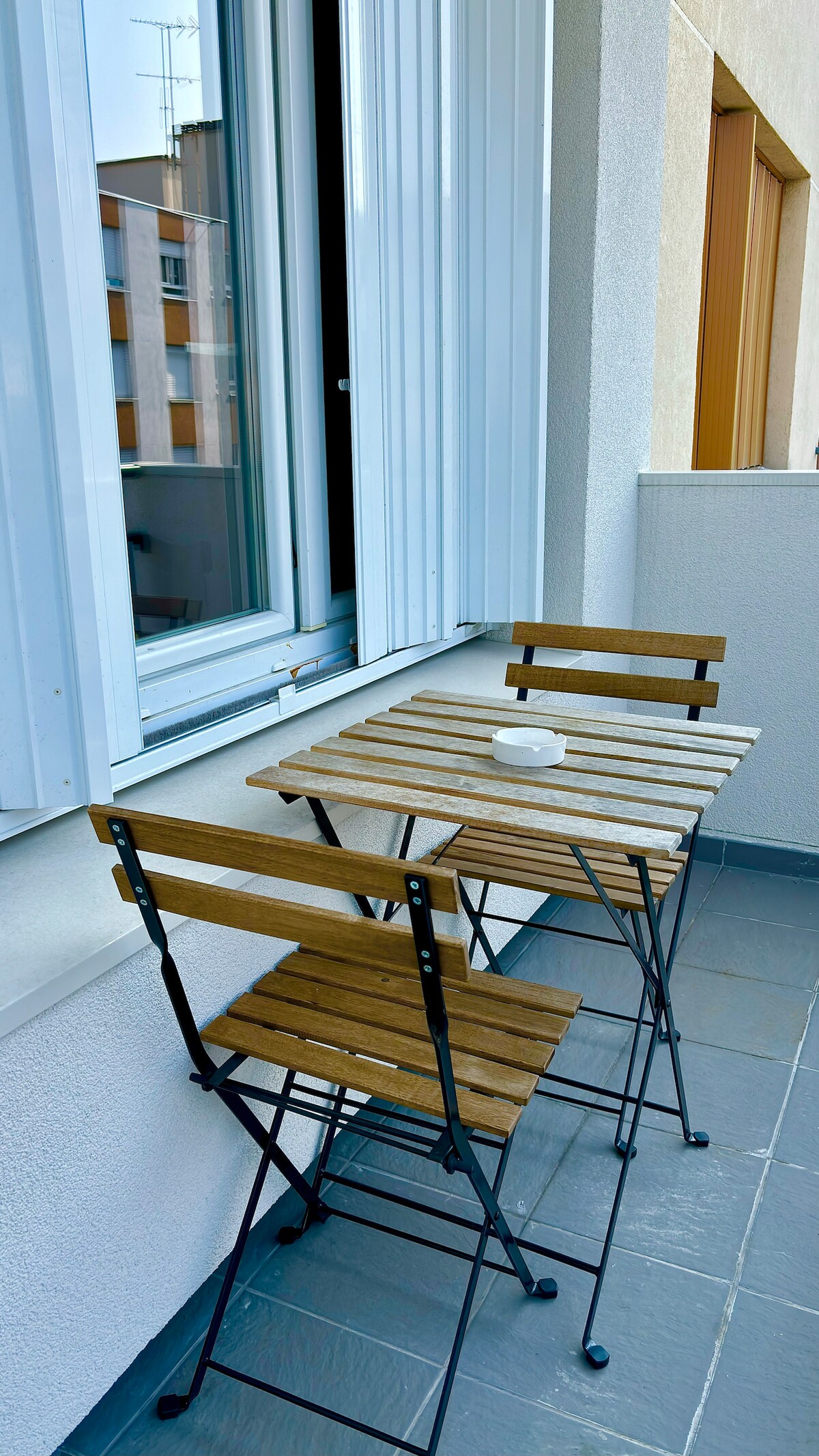 A small balcony features a wooden table accompanied by two folding chairs. The sleek surface is complemented by simple lines and a minimalist design. Light reflects off the neutral-colored walls, and a partial view of the surrounding area is visible through the open window.