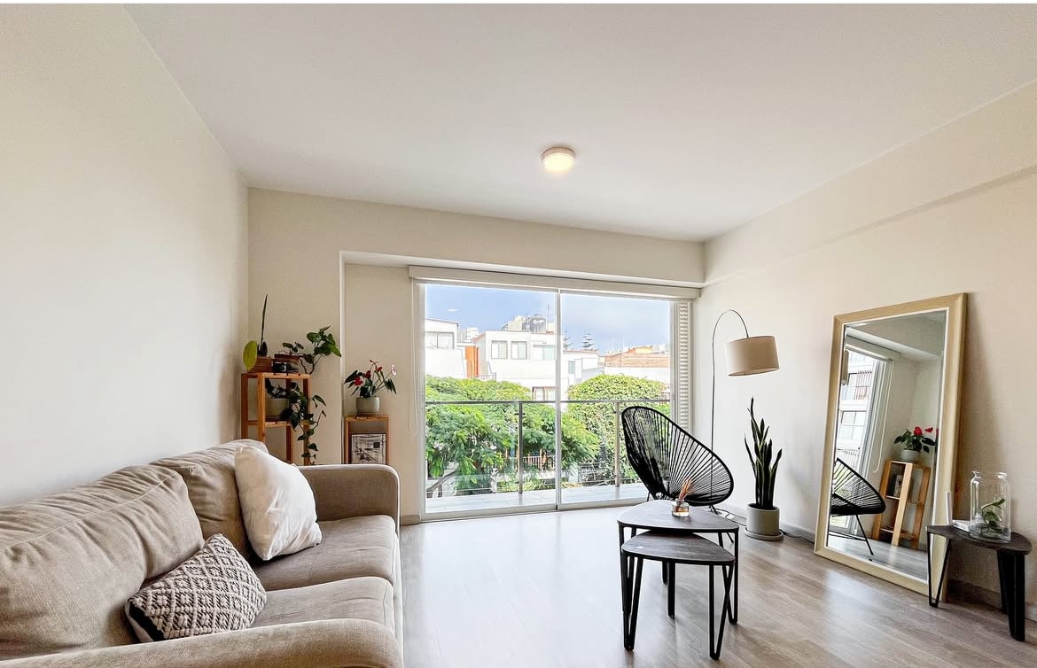 A living area features a light-colored sofa beside a black accent chair and small table. Large glass doors offer access to a balcony overlooking greenery. A full-length mirror and potted plants add to the space, with ample natural light illuminating the room.