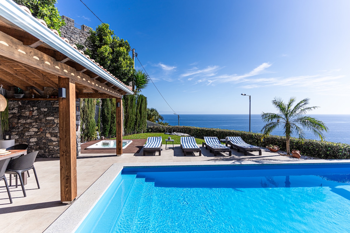 An outdoor space features a clear blue pool with sun loungers arranged around it, overlooking the ocean. A shaded dining area with wooden furniture is positioned nearby, with lush greenery and a palm tree enhancing the scenery. The sky is bright and clear.
