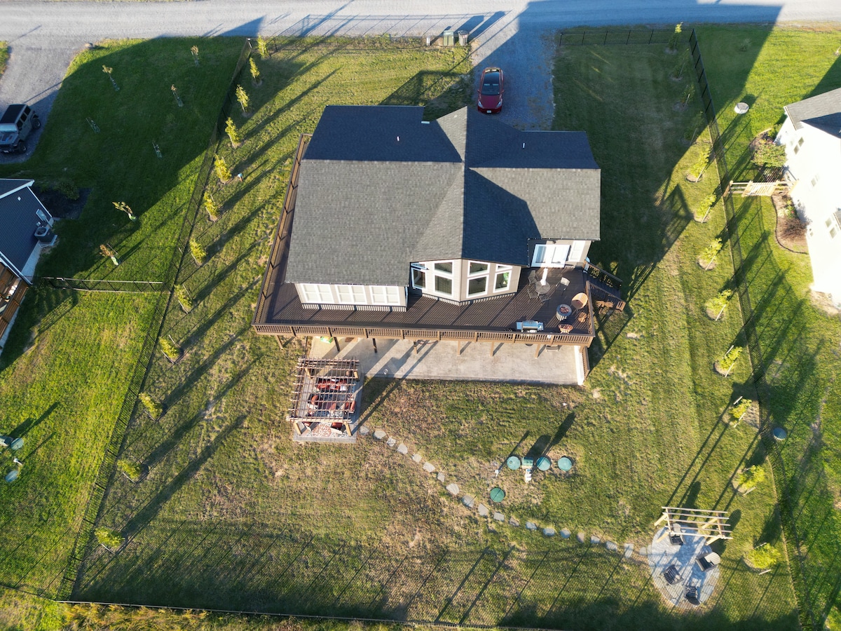 An aerial view of the cozy chalet reveals a spacious deck surrounded by a fenced yard. The green landscape is complemented by evenly spaced shrubs. The structure features multiple windows, and a defined pathway leads to an outdoor seating area.