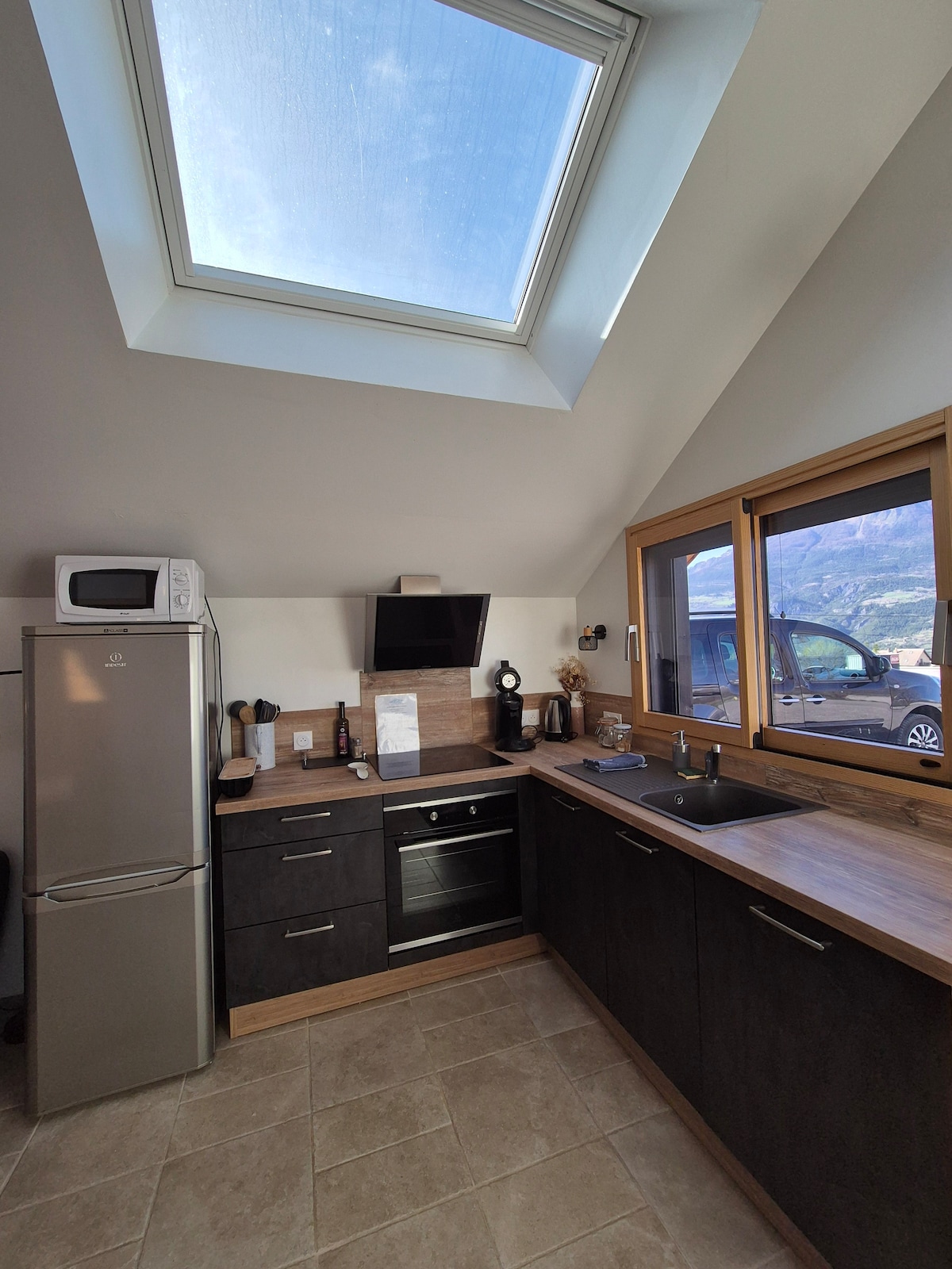 A modern kitchen area is featured, equipped with a refrigerator, oven, and a sink beneath a skylight. Natural light floods the space, highlighting the dark cabinetry and a light-tiled floor. Windows allow views of the surrounding landscape, creating an airy atmosphere.