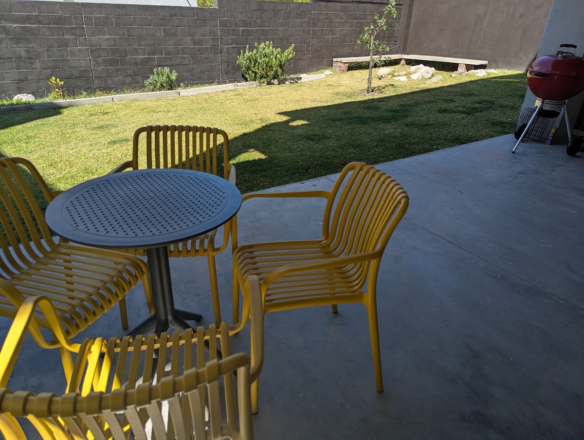 A patio area features a round table surrounded by four yellow chairs. The space includes a visible grassy area with low-maintenance plants in the background. A grill is positioned nearby, and the setting is enhanced by ample natural light.