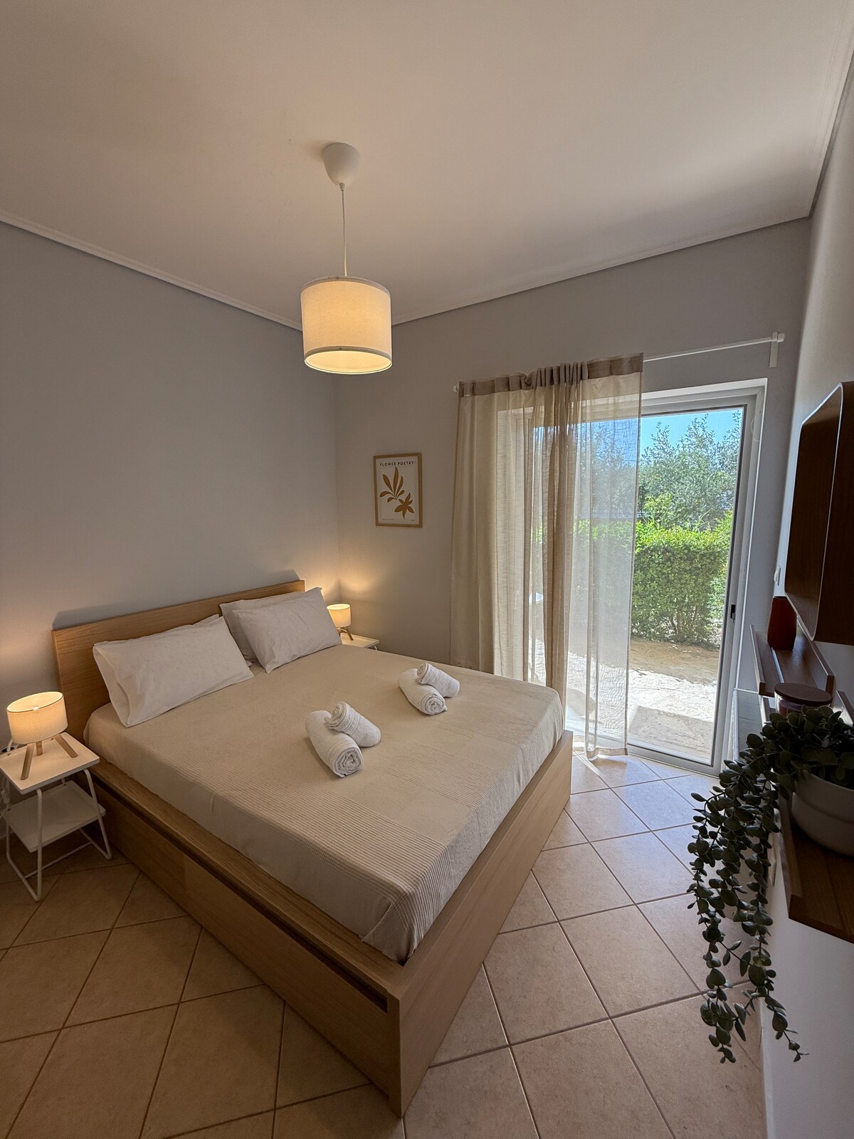 A light-filled bedroom features a spacious bed with soft linens and two rolled towels placed on top. Nightstands with lamps provide soft lighting. A large window with sheer curtains opens to a view of lush greenery, enhancing the room's serene atmosphere.