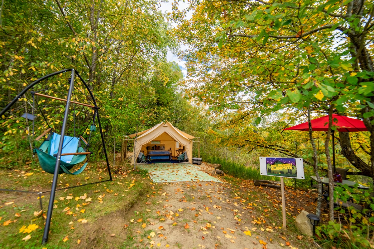 A secluded glampsite features a spacious canvas tent nestled among trees adorned with autumn leaves. A colorful outdoor rug extends from the tent entrance, and a swing chair is positioned nearby. A shaded patio set is visible, enhancing the outdoor relaxation space.