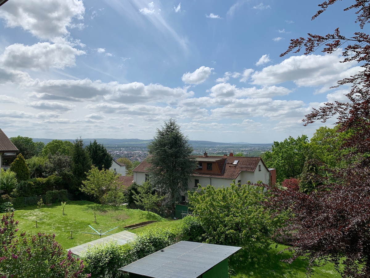 A scenic view presents lush green trees and a sprawling garden, complemented by a serene sky filled with fluffy clouds. The distant landscape showcases rooftops and rolling hills, creating a tranquil outdoor setting perfect for relaxation.