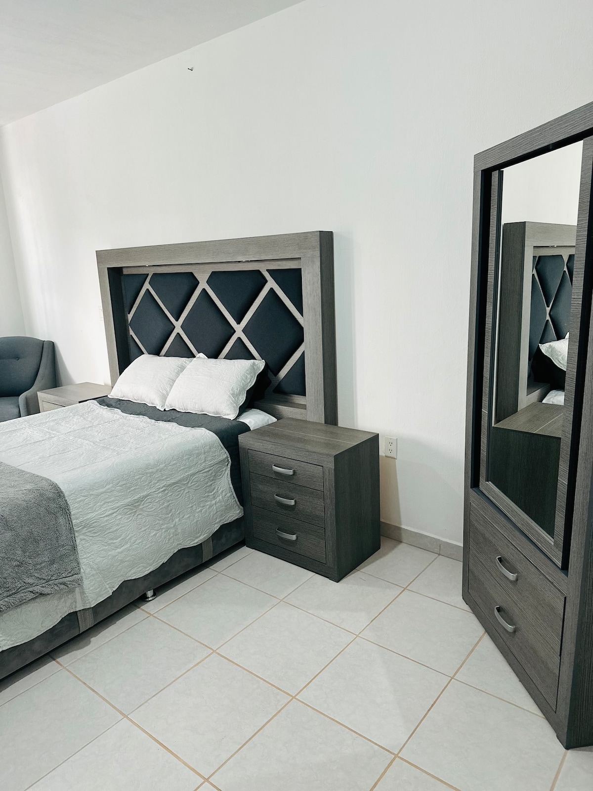 A bedroom features a modern bed with a stylish headboard. A bedside table with drawers is positioned next to the bed. A large mirror is visible, reflecting the room's minimalistic design. A cozy armchair is placed in the corner, enhancing the space's functionality.