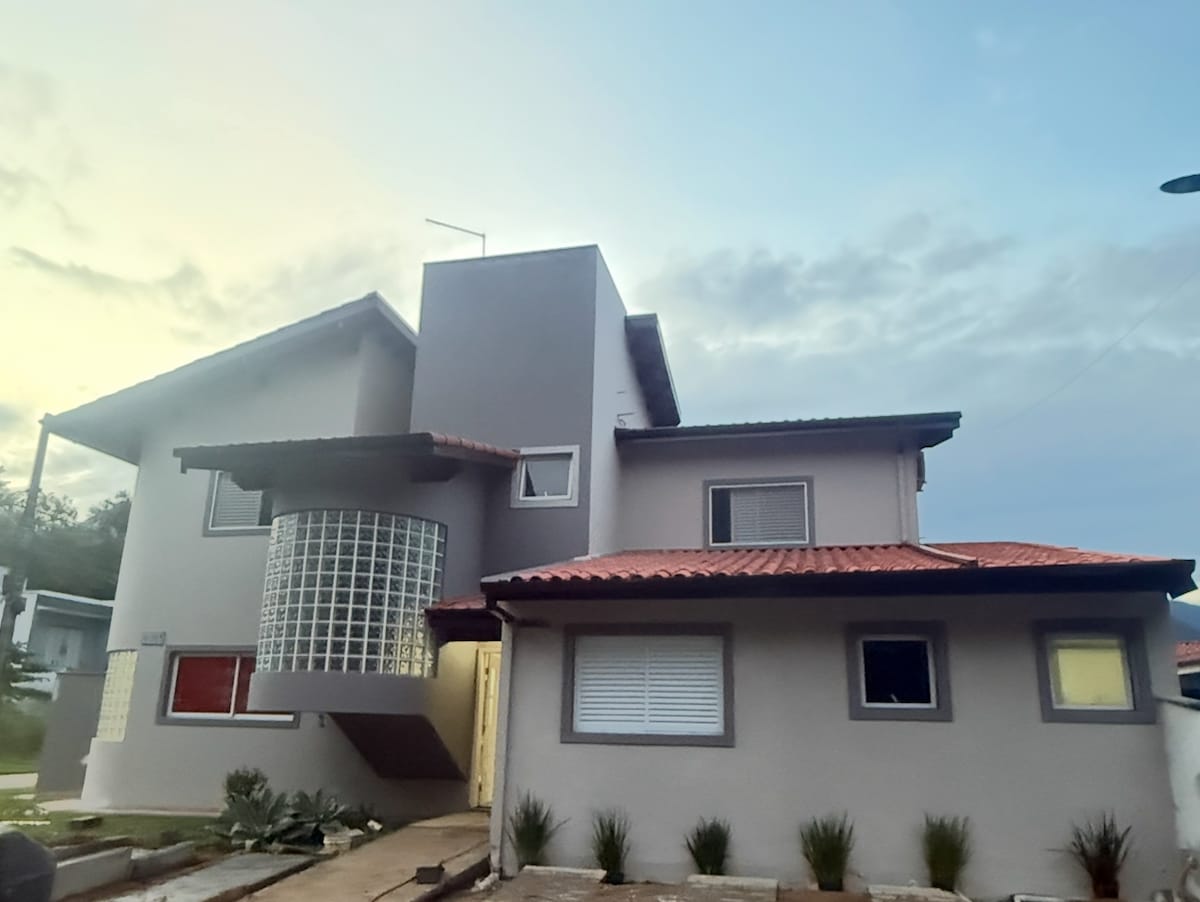 The exterior of a two-story house is shown, featuring a combination of smooth surfaces and textured walls. Large windows allow natural light to enter, while a distinctive glass block wall adds character. A red-tiled roof contrasts with the gray facade, surrounded by neatly landscaped greenery.