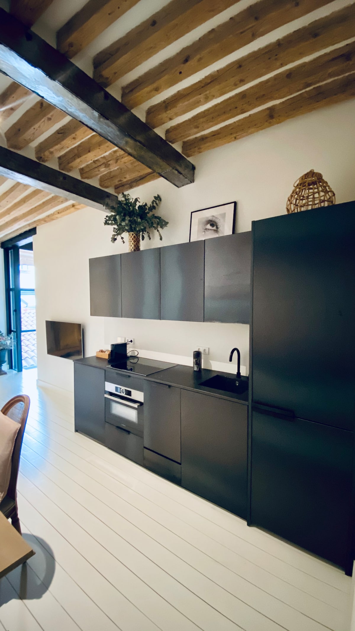 A modern kitchen features sleek black cabinetry and stainless steel appliances, including a built-in oven and refrigerator. A minimalist design is complemented by wooden beams overhead and a decorative plant. A cozy dining area is visible nearby, enhancing the inviting space.