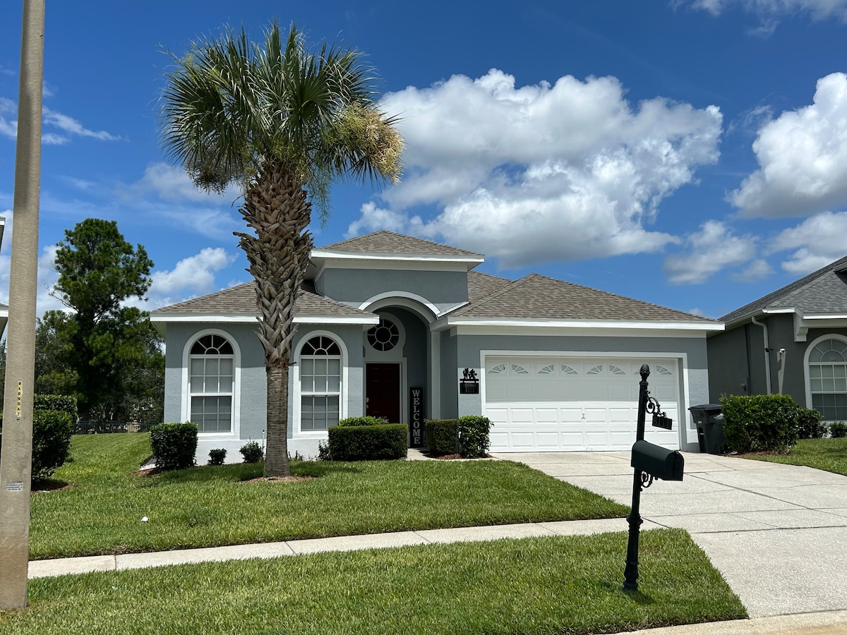 A single-story home features a prominent entrance with arched windows and a welcoming front door. Lush green landscaping surrounds the property, while palm trees sway gently in the foreground. A two-car garage is visible, set against a backdrop of blue skies and puffy clouds.