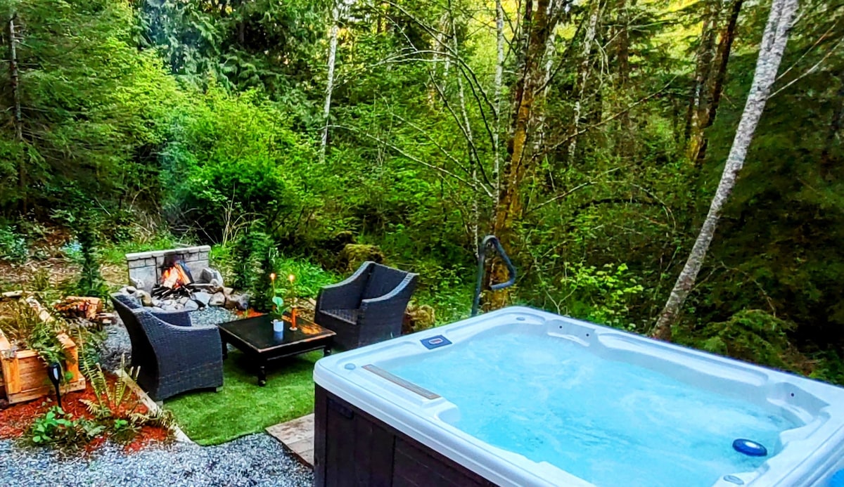 An outdoor hot tub is positioned nearby a stone fire pit, surrounded by lush greenery. Two dark wicker chairs accompany a low coffee table, inviting relaxation amidst the natural setting. The area is softly illuminated, creating a tranquil atmosphere.