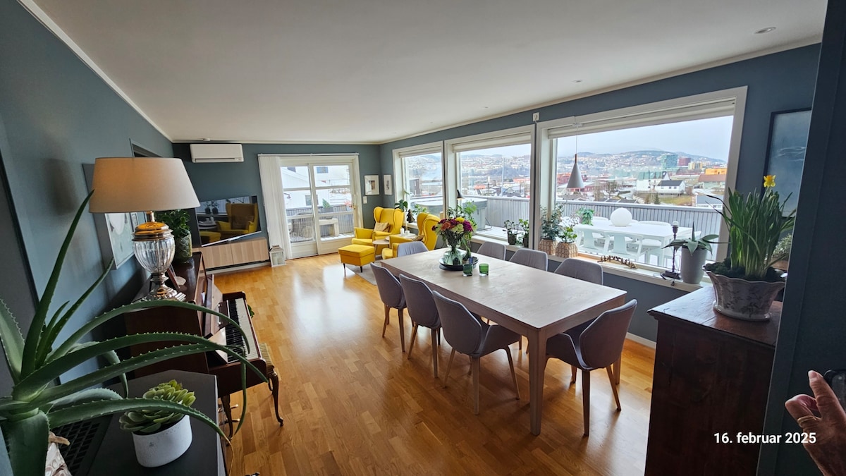 A spacious living area features large windows offering panoramic views. A long dining table is surrounded by modern chairs, while a yellow armchair adds a pop of color. Green plants are placed on surfaces, contributing to a fresh, well-lit environment with wooden flooring.