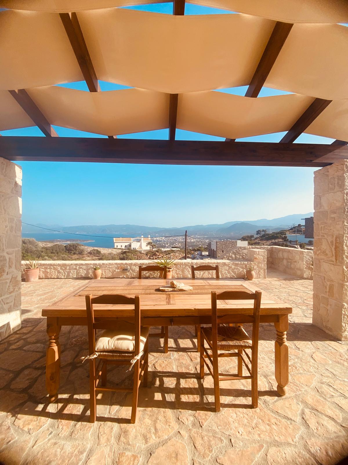 An outdoor dining area features a rustic wooden table surrounded by chairs under a canopy. A panoramic view of the Mediterranean Sea and distant mountains is visible in the background, with the town of Agios Nikolaos partially seen. Natural stone walls frame the inviting space.