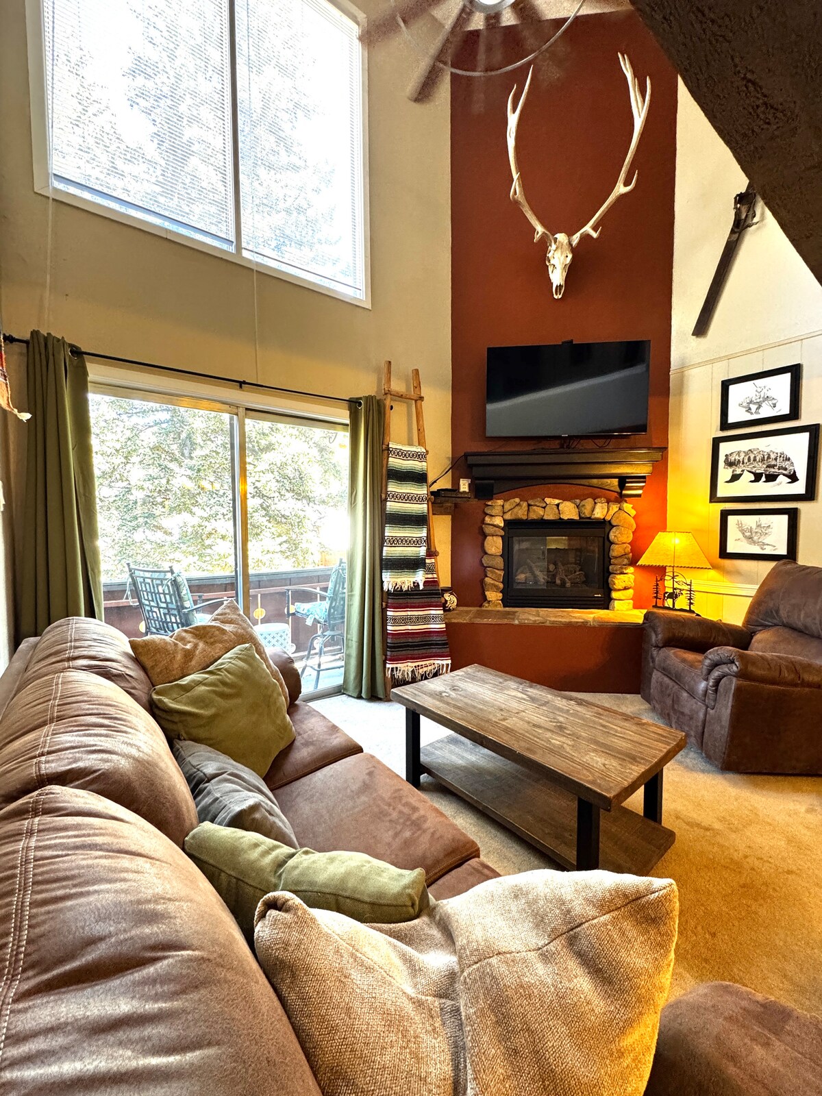 A cozy living area features a gas fireplace surrounded by a stone facade. Large windows invite natural light, while a balcony with outdoor seating is visible. A plush couch with decorative pillows offers comfortable seating, complemented by a wooden coffee table and a mounted television.