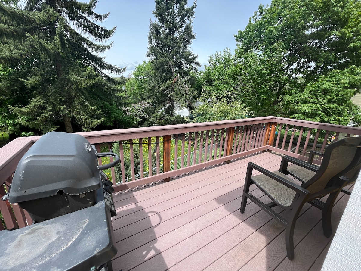 A wooden deck is visible, featuring a charcoal grill and a black chair. Lush greenery surrounds the area, offering a sense of privacy. The view extends to trees and a glimpse of a body of water in the distance, under a clear blue sky.