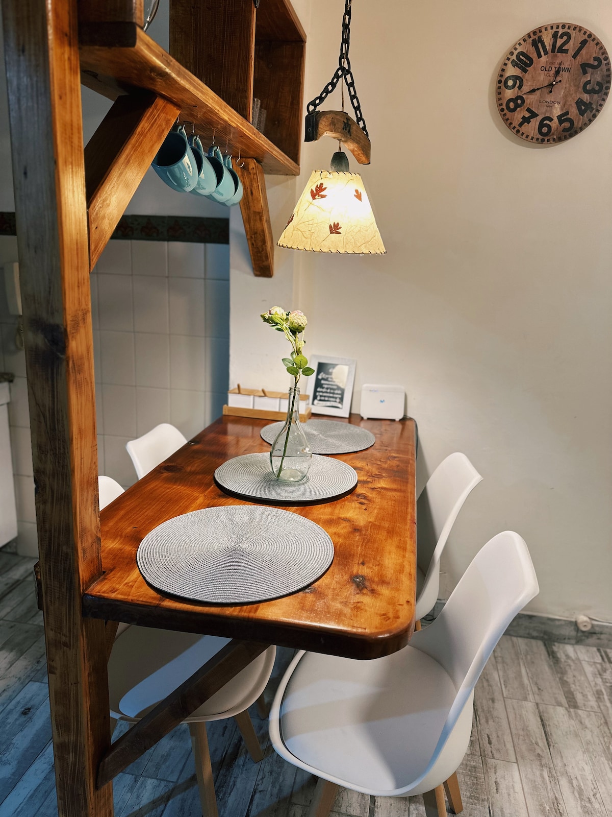 A wooden dining table is set for four with round placemats and a vase containing fresh flowers at its center. White chairs surround the table, and a soft pendant light hangs above. A wall clock is visible in the background.