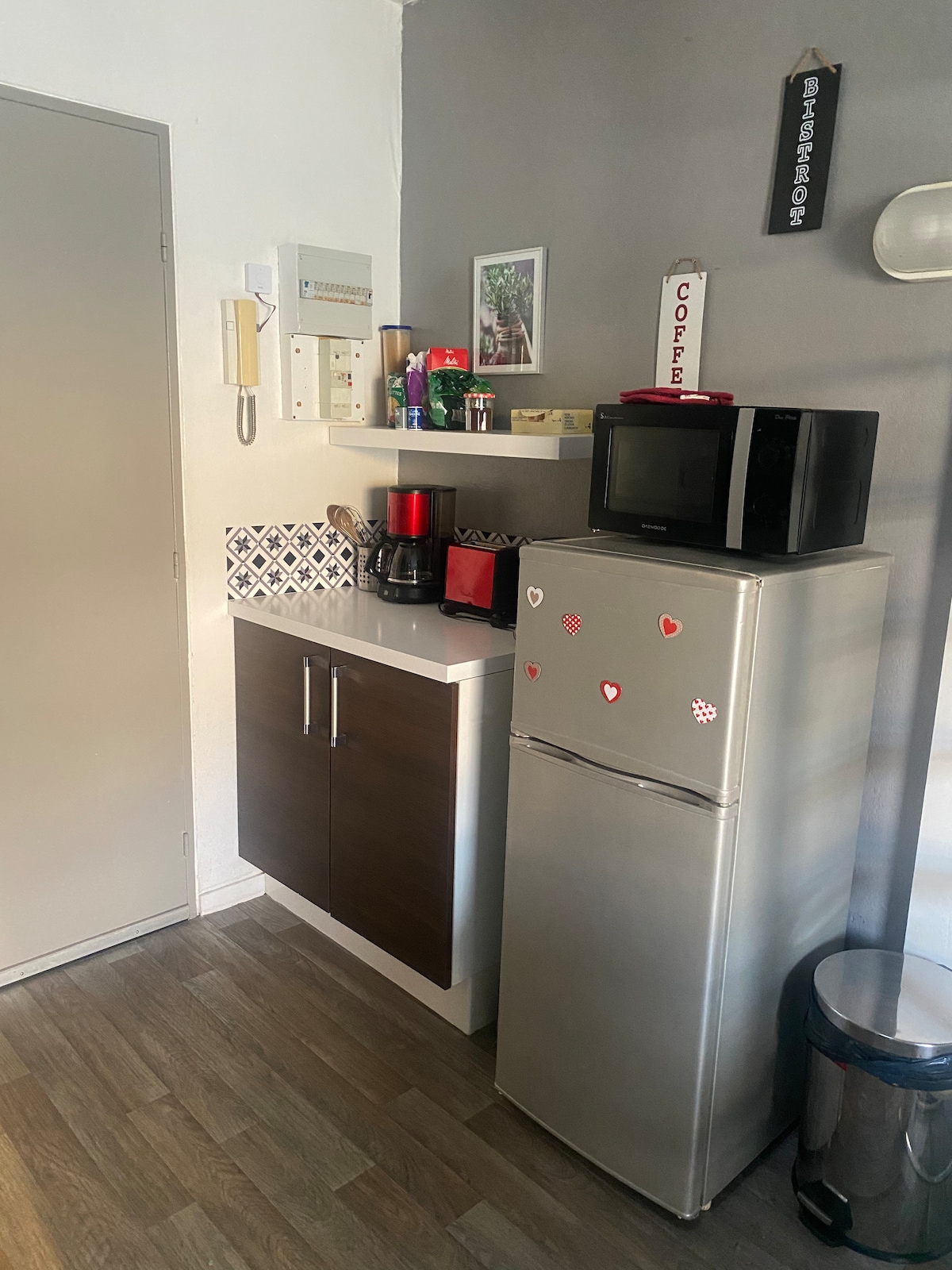 A compact kitchen area features a modern refrigerator with a silver finish and an accompanying microwave on a shelf above. Upper and lower cabinets provide storage, while a coffee machine and utensils can be seen on the countertop, contributing to a functional culinary space.
