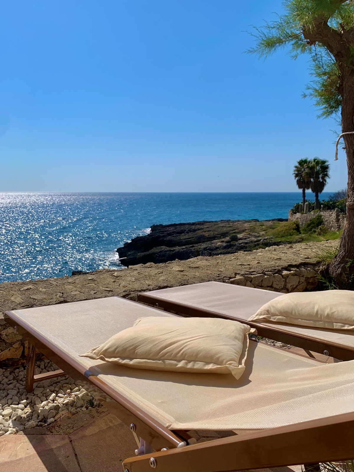 Two lounge chairs are positioned on a terrace overlooking the glittering sea. The coastline features rocky cliffs, and palm trees are visible in the background, adding to the serene atmosphere. The deep blue water reflects sunlight, creating a tranquil setting.