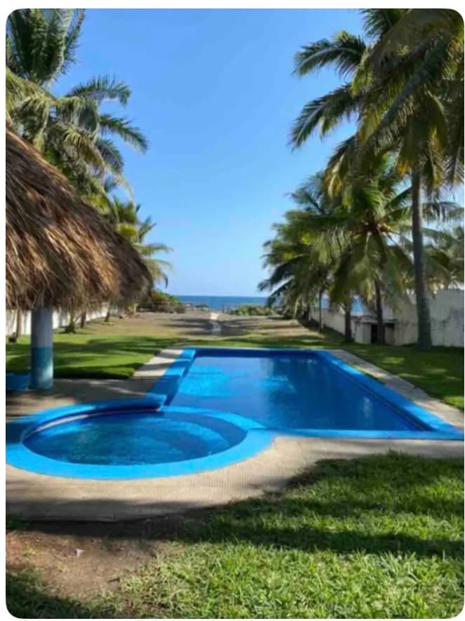 A spacious outdoor area features a large pool and a separate jacuzzi, both surrounded by a manicured lawn with tropical palm trees. The ocean is visible in the distance, providing a serene backdrop. A thatched-roof gazebo offers shaded seating nearby.