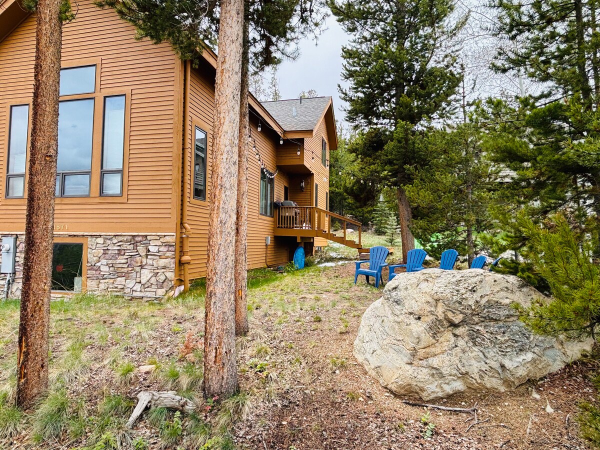 The exterior of a spacious home is shown, framed by tall pine trees. A seating area with blue chairs is visible on the deck, while a large rock and green foliage provide natural elements to the landscape.