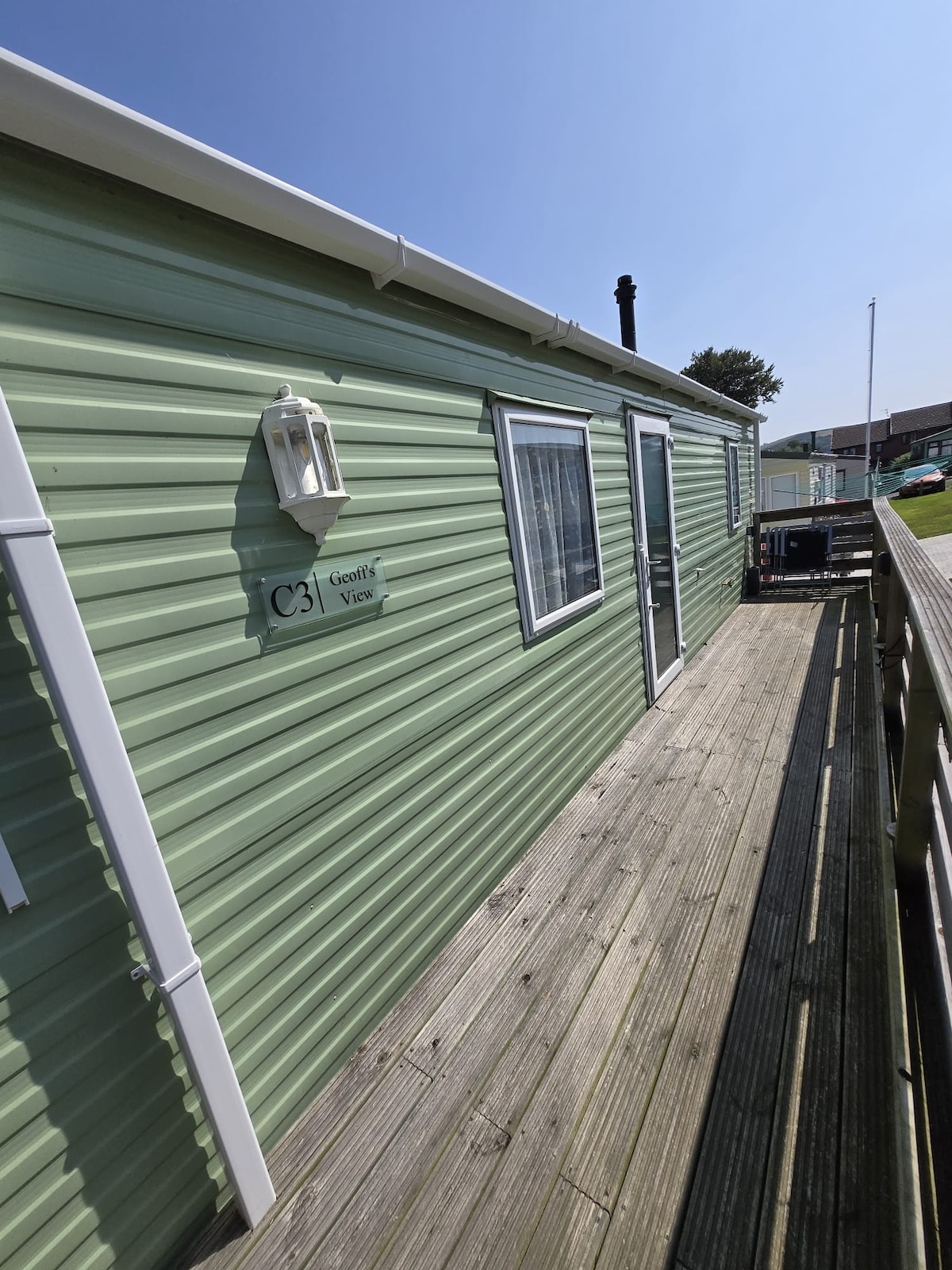 The exterior of the caravan is shown, featuring green siding and a wooden deck. A sign indicating 'Geoff's View' is mounted on the wall. The entrance is visible with a nearby window, and the clear sky above suggests bright weather.