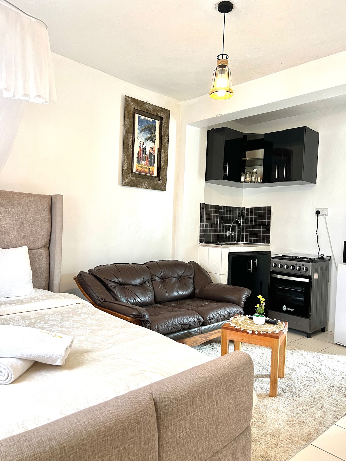 A cozy space is shown, featuring a spacious bed with crisp white linens and a soft, inviting headboard. A comfortable brown leather loveseat complements the area, with a small wooden coffee table. A compact kitchenette is visible in the background, equipped with essential appliances.