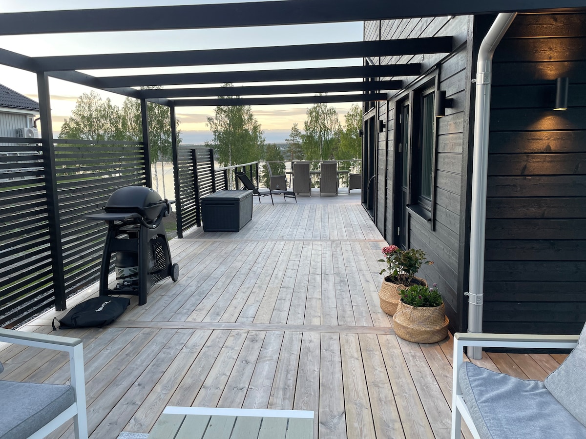 A spacious wooden terrace features a gas grill and comfortable seating. Potted plants add a touch of greenery, while views of the surrounding nature are visible in the background. The area is designed for relaxation and outdoor dining.