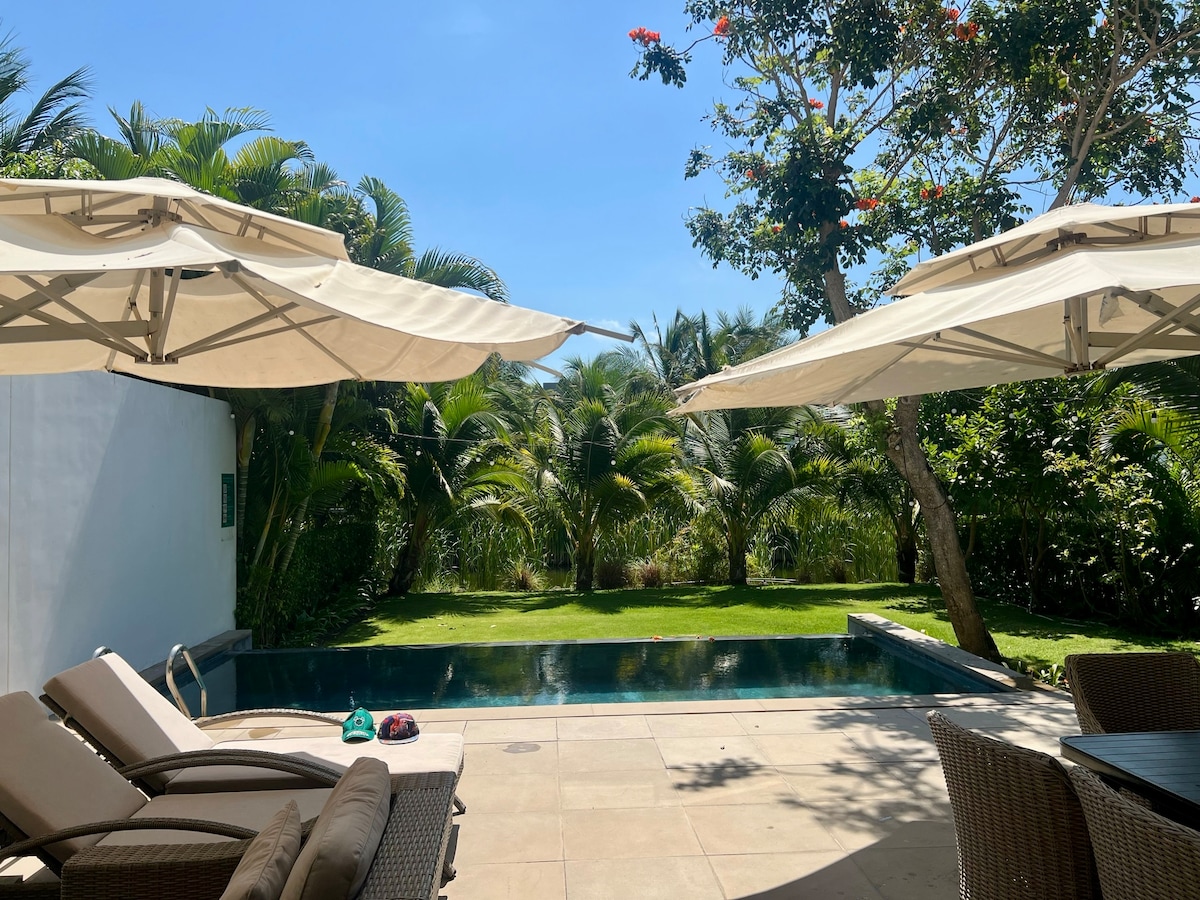 The outdoor area features a tranquil swimming pool surrounded by lush greenery. Comfortable lounge chairs are positioned under large umbrellas, providing shade. The serene setting is complemented by tropical plants and a clear blue sky, creating a relaxed atmosphere for enjoyment.