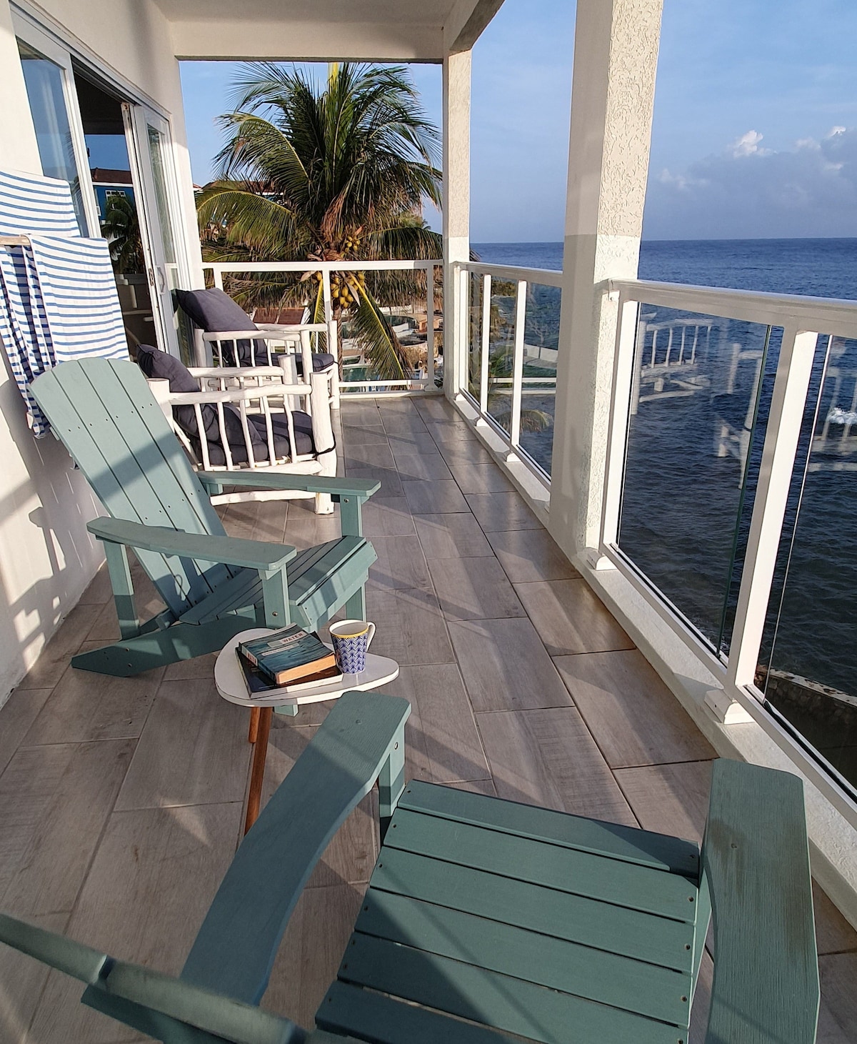 A verandah offers a serene space with two comfortable chairs and a small table. A stack of books rests on the table beside a cup, with views of the ocean and palm trees in the background, enhancing a peaceful atmosphere.