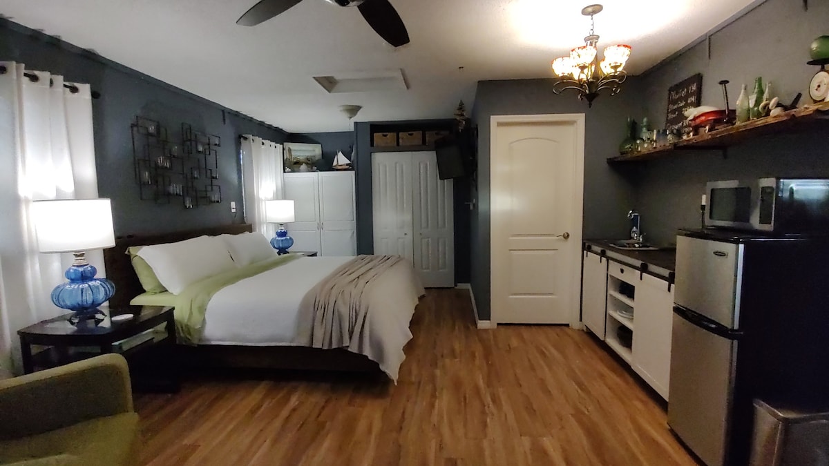 The guest suite features a spacious bed with soft linens and an accent pillow, flanked by two lamps on bedside tables. A kitchenette with modern appliances is visible, along with a cozy green lounge chair in the corner. Natural light enters through sheer window treatments.
