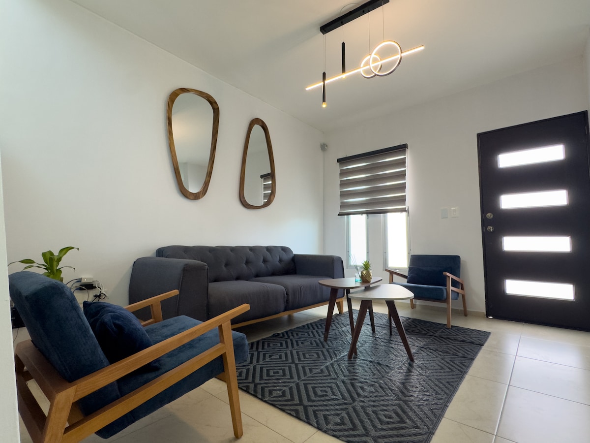 A modern living area is highlighted by a comfortable gray sofa and two wooden armchairs. A round coffee table sits at the center, complemented by a subtle plant. Natural light is allowed through the window covered with stylish shades, while a contemporary light fixture adds a sleek touch.