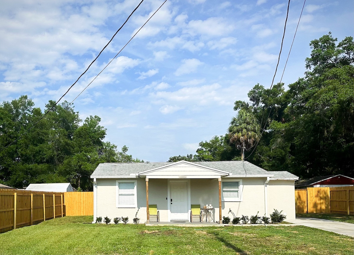The exterior of a remodeled home is presented, featuring a front porch with two chairs. A neatly manicured lawn extends toward a fenced yard. Tall trees and green shrubs provide a natural backdrop, while a clear sky is visible above the home.