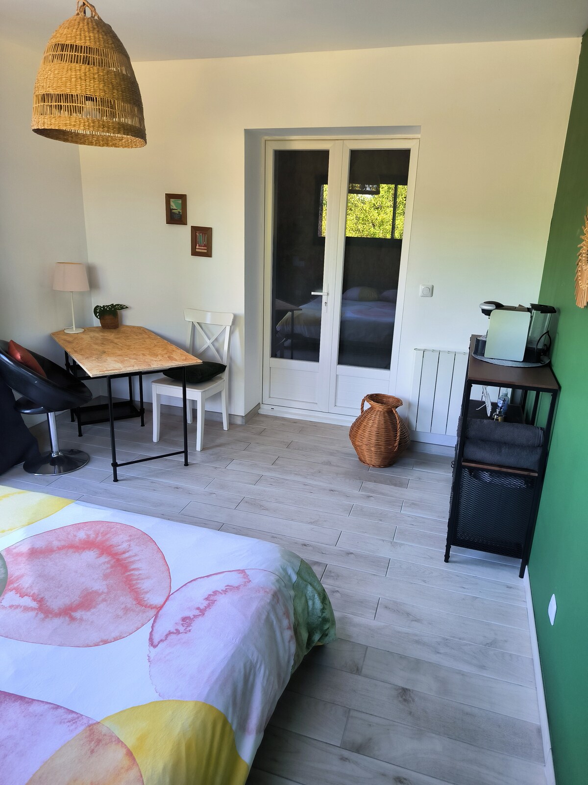 A semi-independent room features a spacious bed with a colorful floral duvet and a small table with chairs. A large window allows natural light, illuminating the modern décor. A wicker pendant light complements the room's simple furnishings, while a small kitchenette and a decorative basket are visible.