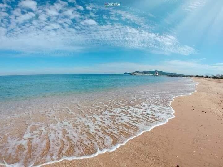 A serene coastal scene captures a stretch of sandy beach meeting clear blue waters. Gentle waves lap at the shore under a bright sky with scattered clouds, creating a peaceful atmosphere. Distant hills and structures are visible along the coastline.