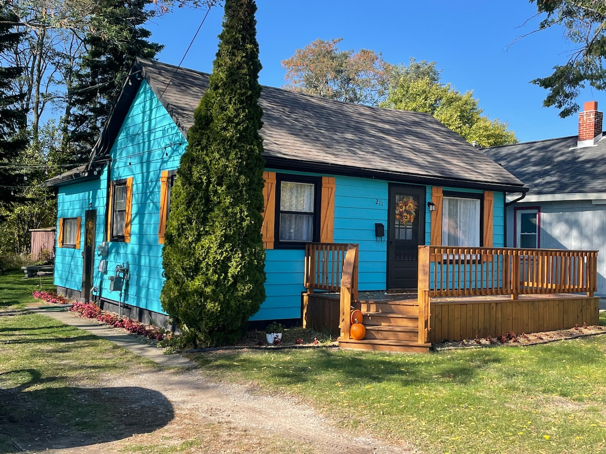 A vibrant blue house features wooden shutters and a welcoming front porch. The porch is bordered by a wooden railing and adorned with seasonal decor. A manicured lawn with flower beds complements the cheerful exterior, while trees provide shade in the background.