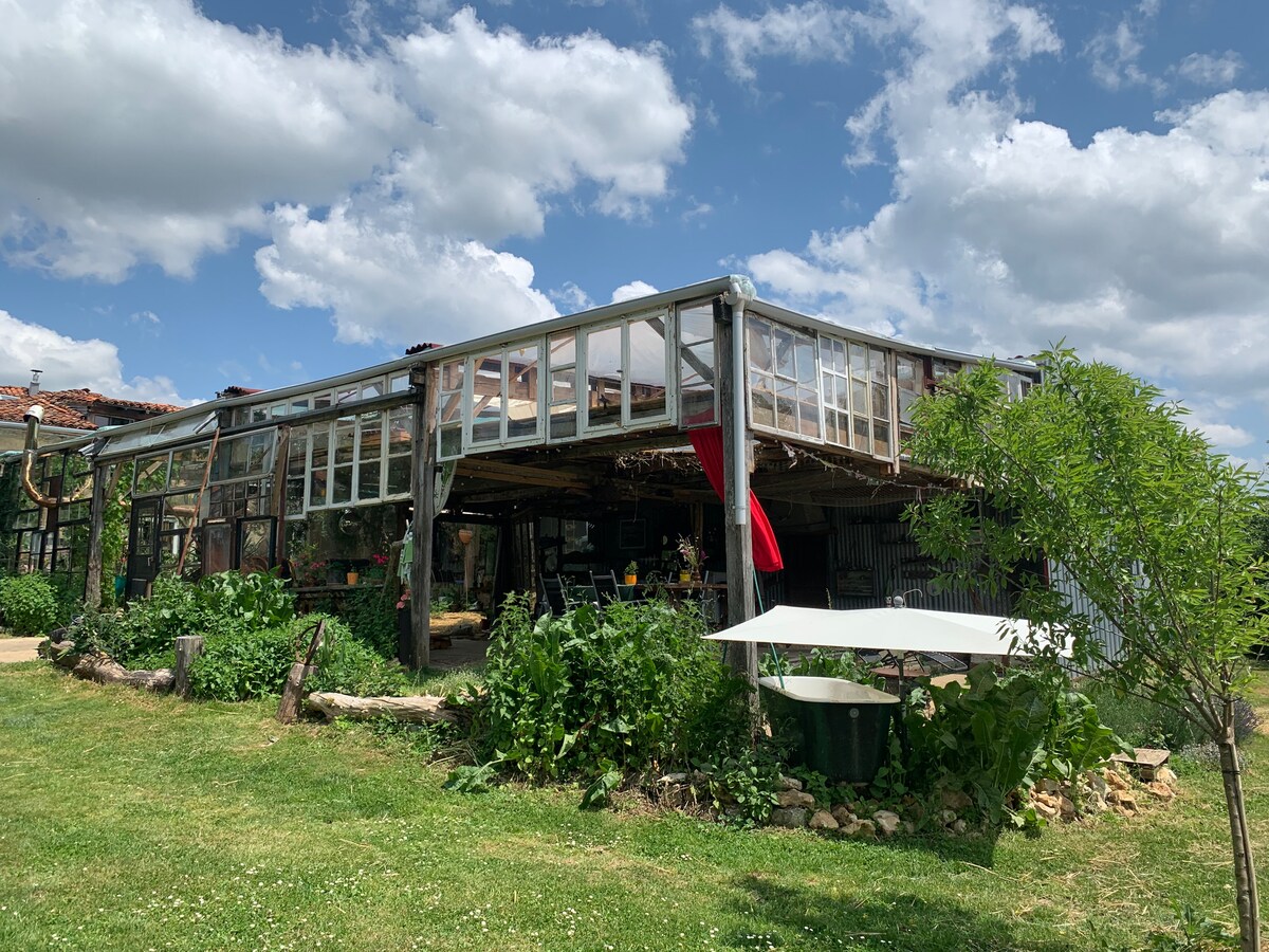A large greenhouse structure is featured, with glass windows and wooden beams revealing a vibrant garden. A white table sits under a shaded area, surrounded by lush greenery. The sky shows scattered clouds, enhancing the outdoor ambiance.