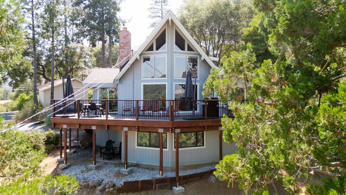 The exterior of Chalet 186 showcases a two-story design with large windows that allow for ample natural light. A spacious deck surrounds the home, furnished with seating for relaxation. Surrounding trees provide greenery, enhancing the home's secluded, mountain retreat feel.