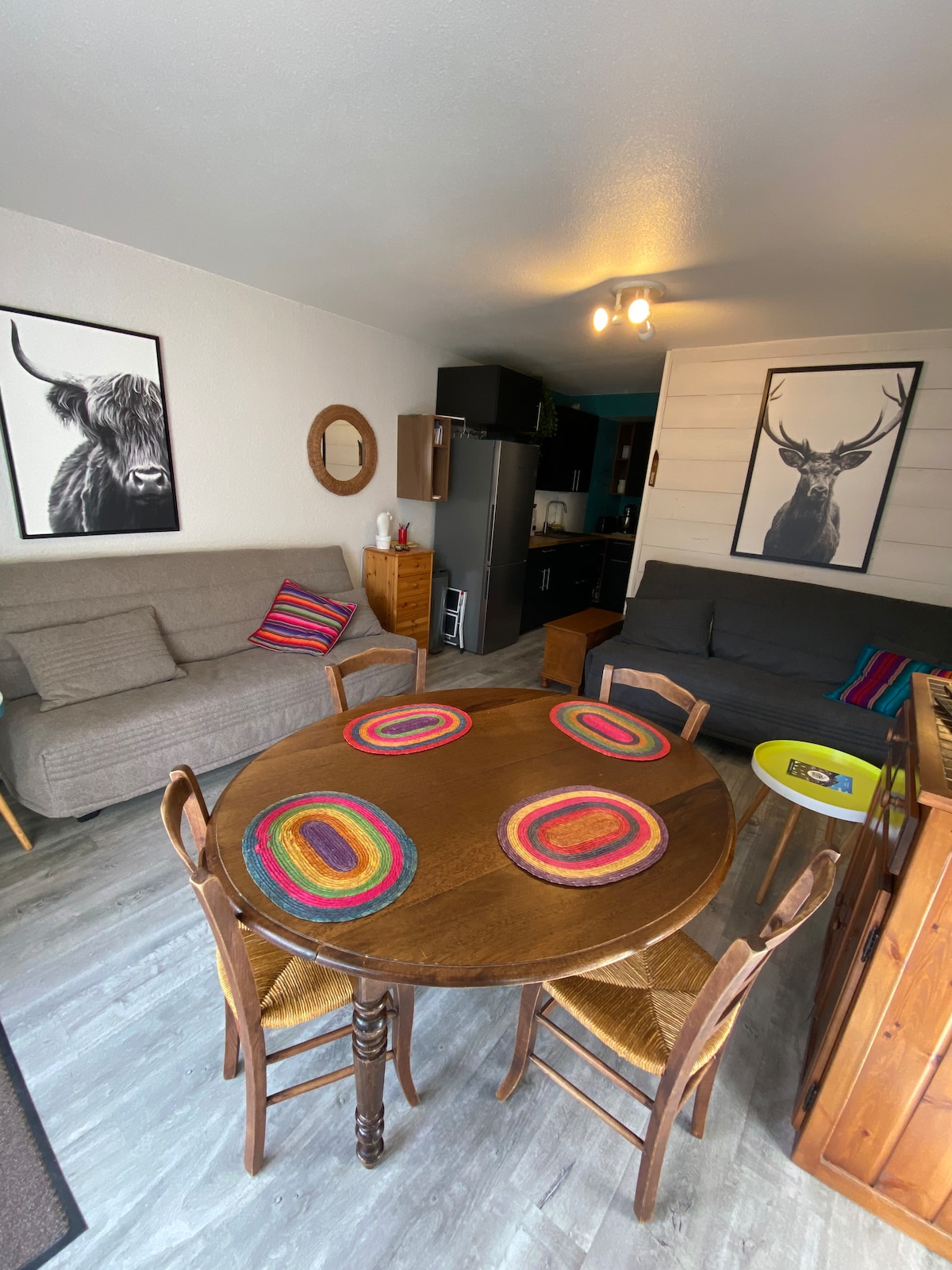 A living area is shown, featuring a round wooden table with colorful placemats. Two chairs are positioned around the table, and a pull-out sofa bed is present against the wall. Art prints of wildlife adorn the walls, and a kitchenette is visible in the background.