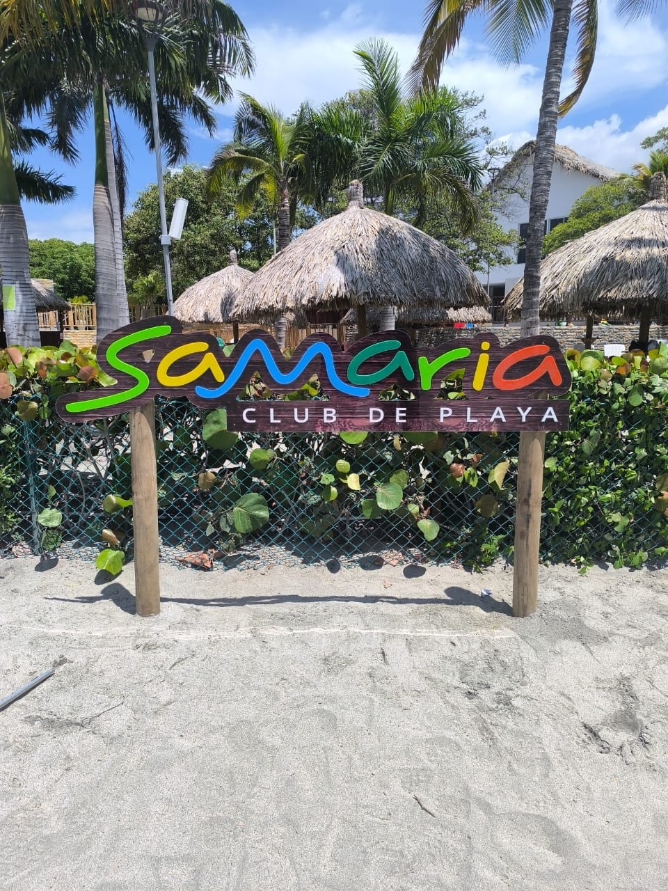 The image depicts the entrance sign for Samaria Club de Playa, featuring vibrant colors against a natural backdrop. Surrounding greenery and palm trees enhance the welcoming atmosphere. Thatched-roof kiosks are visible in the background, indicating a beachside setting with sandy pathways.