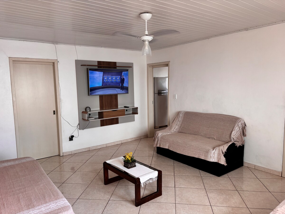 A living area features a flat-screen television mounted on a wall, with a light-colored sofa and a dark sofa adorned with a beige throw. A small coffee table sits in the center, accompanied by a decorative plant, with tile flooring throughout the space.