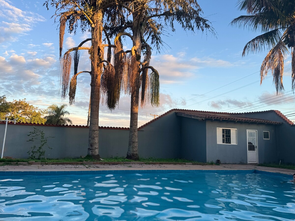 A swimming pool is visible in the foreground, reflecting the clear sky. Tropical palm trees frame the scene, while a modest gray structure with a white door and window is set against a simple outdoor backdrop. The setting offers a serene space for relaxation.