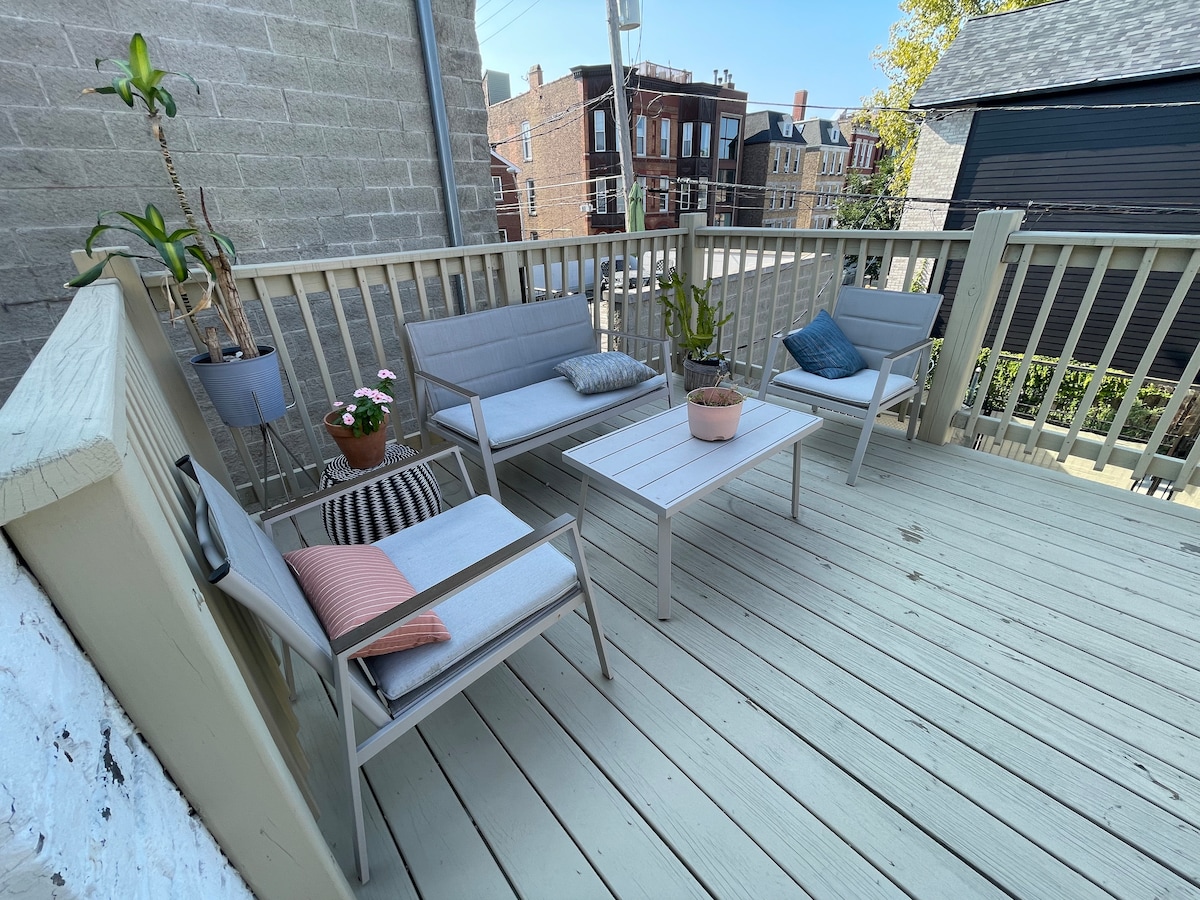 An outdoor deck is furnished with a table and four light gray chairs. Soft cushions are placed on two chairs, and a potted plant sits at the center. The space is surrounded by wooden railings, with nearby buildings visible in the background.