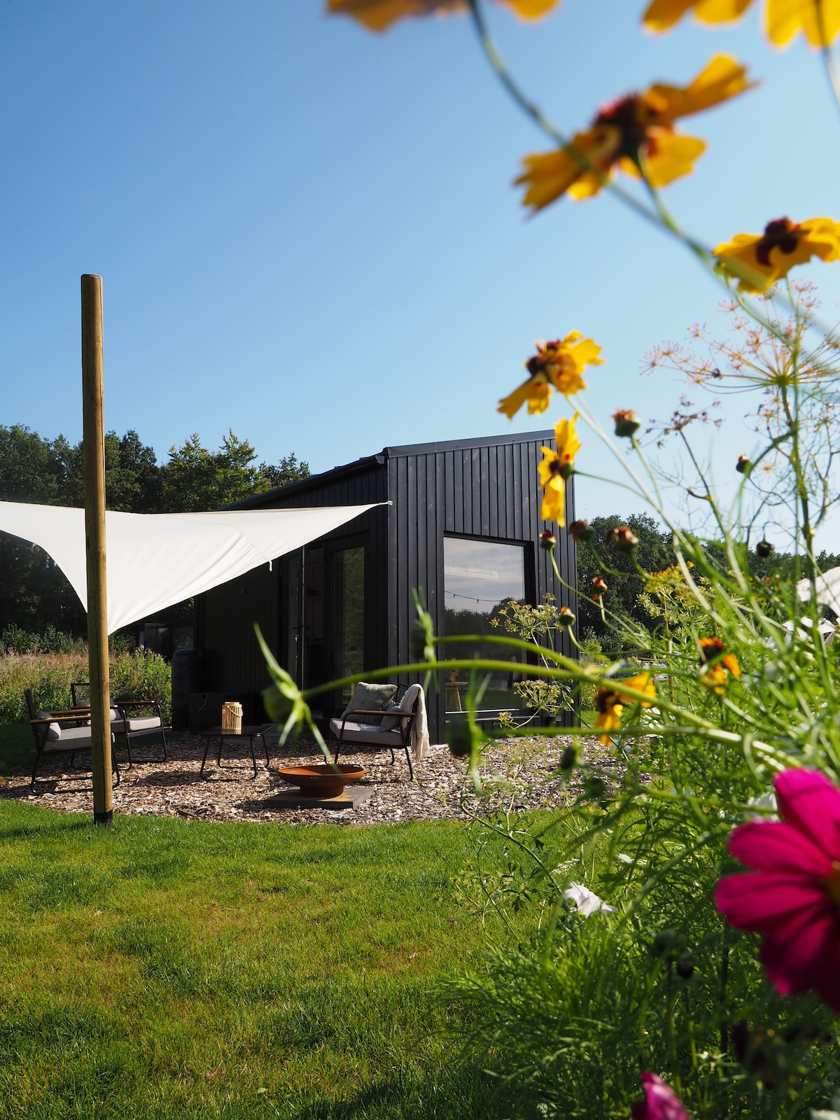 A unique tiny house is positioned amid greenery, featuring a dark exterior and large windows. A lounge set rests on a pebbled patio, under bright sunlit fabric overhead. Flowers in vibrant hues surround the space, adding natural color to the tranquil setting.