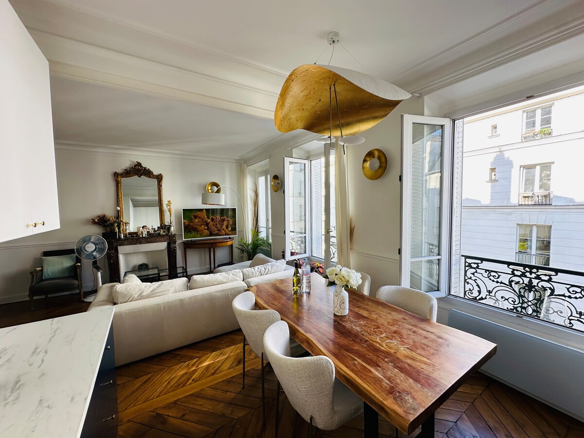 A dining area features a large wooden table surrounded by upholstered chairs. Natural light fills the space through double windows, highlighting the elegant decor. A cozy seating area is visible, with a TV positioned for easy viewing and a decorative fan adding to the ambiance.