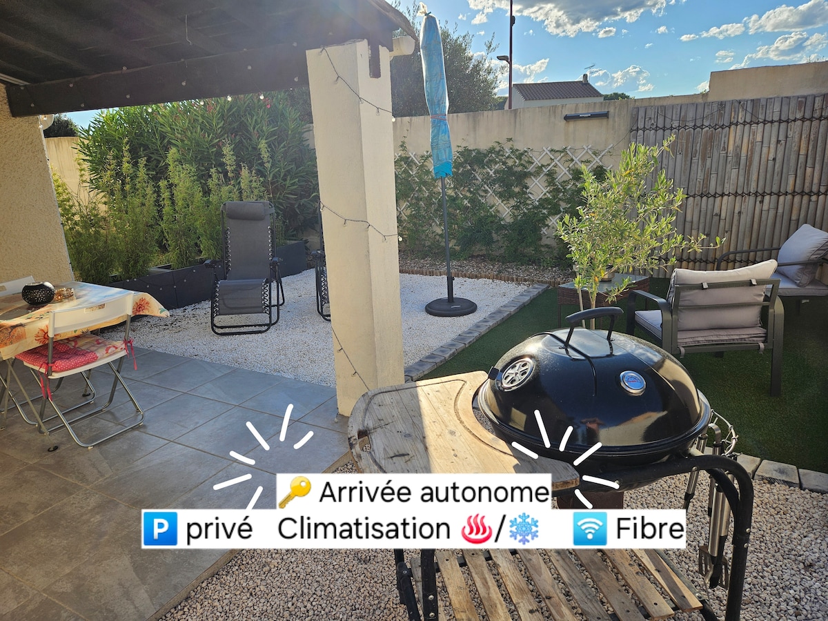 An outdoor area is featured with a charcoal barbecue, a small dining table, and comfortable seating arrangements. The space is surrounded by greenery and a wooden fence, providing privacy. Soft lighting options and a shaded area contribute to a cozy ambiance.