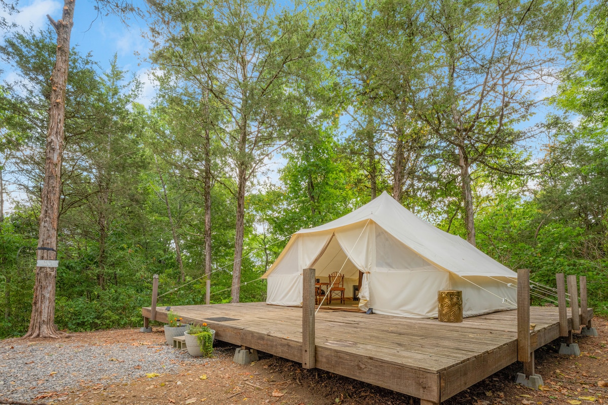 A spacious glamping tent is set on a wooden deck surrounded by dense greenery. The canvas structure features an inviting entrance, revealing a cozy seating area inside. Potted plants decorate the deck, enhancing the natural atmosphere of the site.