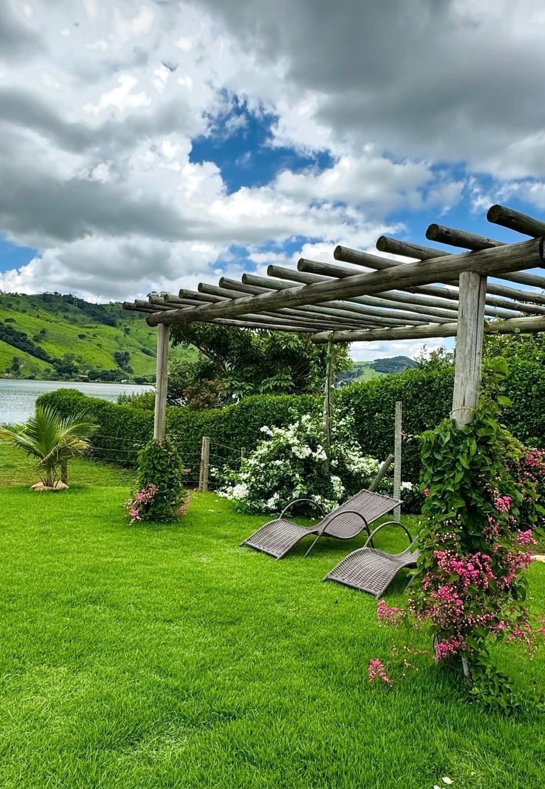 A spacious outdoor area is highlighted by a wooden pergola, providing shade over two lounge chairs. Lush greenery surrounds the space, complemented by flowering plants in shades of pink and white. A tranquil lagoon is visible in the background under a cloudy sky.