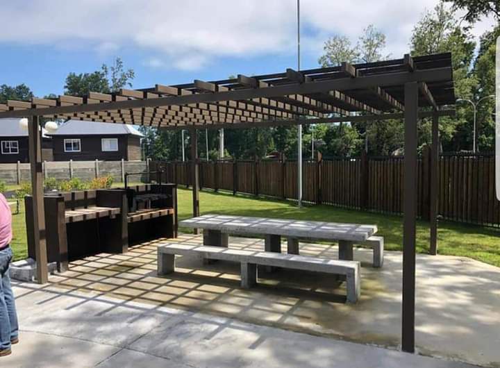 An outdoor gathering area features a shaded pergola supported by a wooden frame. Underneath, a spacious table is accompanied by several benches. The concrete surface provides a durable setting, while grass surrounds the area, promoting a natural atmosphere for relaxation.