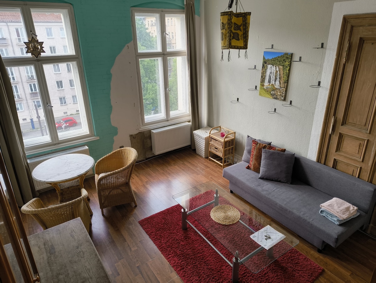 A bright living area is presented, featuring a grey sofa and a glass coffee table on a red rug. Natural light floods in through large windows, with light green walls enhancing the space. A small dining table with wicker chairs and wall-mounted shelves are also visible.