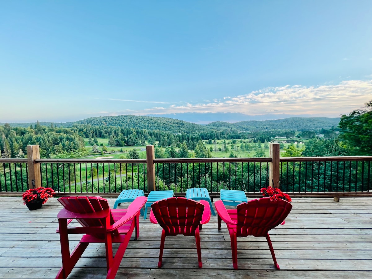 A spacious outdoor deck features bright red Adirondack chairs and matching side tables, offering a clear view of rolling green hills and mountains in the distance. Lush trees surround the area, enhancing the serene setting with a touch of nature.