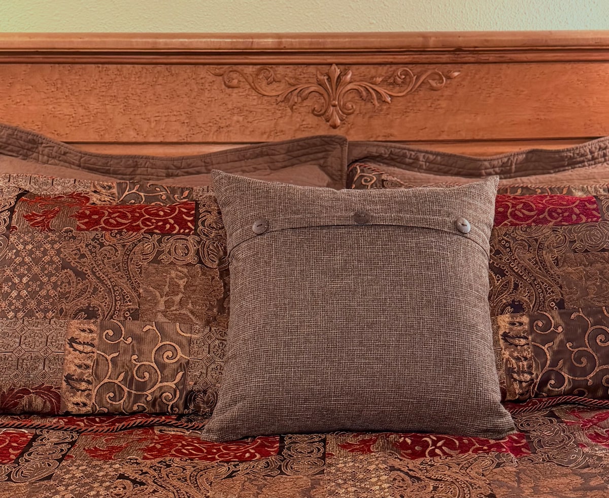 The image highlights a beautifully adorned bed with rich, textured bedding featuring intricate patterns in warm tones. A decorative, buttoned throw pillow sits prominently in the center, adding an inviting touch to the overall arrangement.